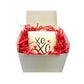 A white gift box with red shredded paper holds the XOXO Colored Quote Candle in a frosted tumbler jar, featuring a label with "XOXO" and small red hearts.